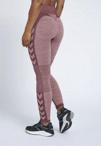 Leggings di un colore bordeaux con maglia testurizzata e pannelli in rete. Presentano dettagli a chevron lungo il lato. Indossati con scarpe da ginnastica nere.