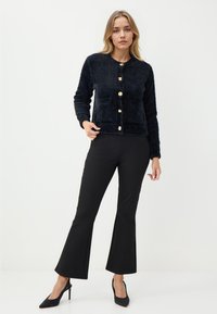 Cardigan nero con una texture pelosa e bottoni dorati, abbinato a pantaloni neri a palazzo e tacchi neri, sullo sfondo neutro.