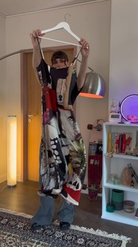 Bunt gemustertes Kleid aus leichtem Stoff, das verschiedene Drucke in Schwarz, Rot und Beige zeigt. Das Modell hält es an einem weißen Kleiderbügel.