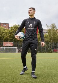 Black athletic tracksuit with red accents, featuring a high collar and zip closure. A soccer ball is held in the left hand. Black sport shoes.