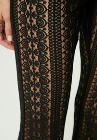 Leggings in pizzo nero con dettagliati motivi floreali e geometrici, caratterizzati da una texture trasparente e elementi di design ornate lungo i lati.