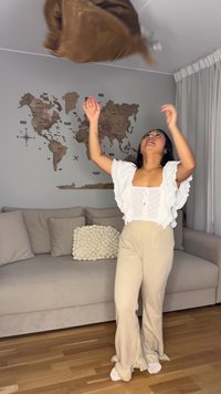 Lätt brun väska kastas. Personen bär en vit texturerad topp med volangärmar och beige ribbstickade vida byxor.