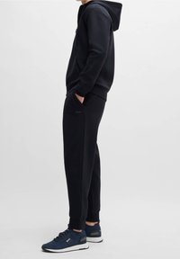 Svart hoodie med ficka fram och matchande joggingsbyxor. Texturerat tyg med åtsittande mudd. Kombinerad med marinblå löparskor.