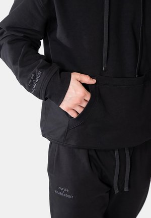 Persoon draagt zwarte hoodie en bijpassende broek met de tekst "nur die x Kilian Kerner," hand in de voorzak van de hoodie.