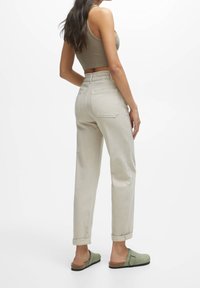 Ljust beige högmidjade byxor med elastisk midja, raka ben och upprullade ärmar, parat med gröna slip-on klacker.