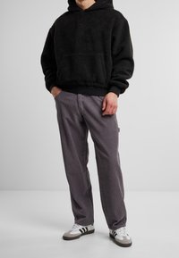 Felpa nera con cappuccio e tasca a marsupio, abbinata a pantaloni grigi di velluto a coste. Il modello indossa scarpe da ginnastica bianche con strisce nere.