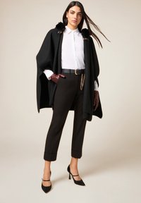 Capa negra con collar de piel sintética, camisa blanca, pantalones marrones y zapatos negros de tacón con correas en el tobillo. Los accesorios incluyen un cinturón de cadena y guantes.