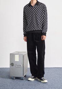 Pull-over à motif noir et blanc avec un col, associé à un pantalon en velours côtelé noir. Près d'une valise trolley métallique argentée sur roulettes.