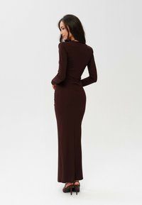 Burgundské dlouhé šaty s těsným střihem a hladkou texturou, končící u kotníků, doplněné o vysoké podpatky.