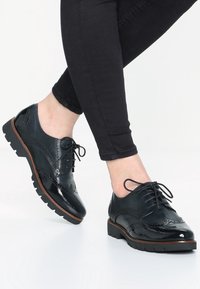 Scarpe oxford in vernice nera con lacci, decorazione brogue e suole robuste, abbinate a jeans neri.