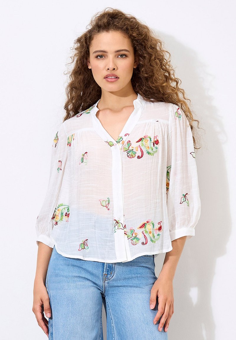 White, lightweight blouse with colorful floral embroidery, v-neckline, and three-quarter sleeves, worn over blue jeans.