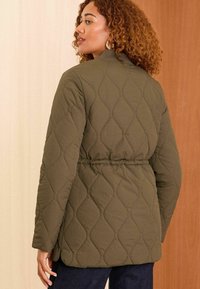 Olīvu zaļš quilted jaka ar jostu viduklī; iezīmē augstu apkakli un teksturētu dimanta rakstu. Aizmugures skats rāda gludu audumu un aproces.