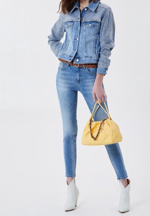 Vrouw in een blauwe spijkerjack met rhinestonedetails, skinny jeans, witte enkellaarsjes en met een gele handtas met kettinghandvatten.