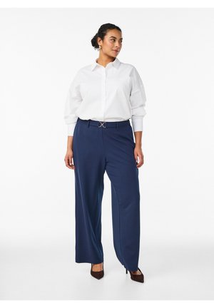 Femme debout portant une chemise blanche à manches longues rentrée dans un pantalon large bleu marine avec une ceinture noire et des escarpins sombres à bouts pointus.