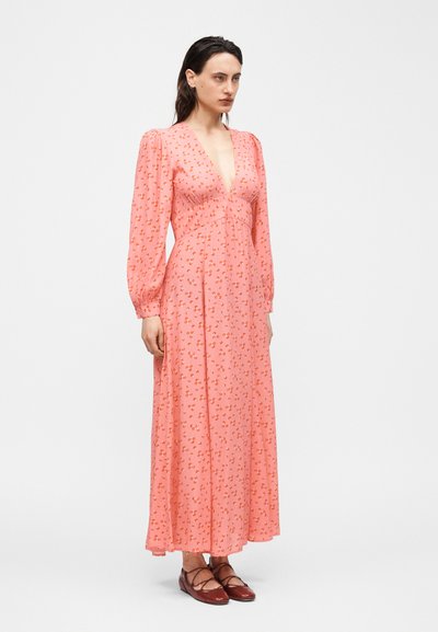 Γυναίκα που φοράει μακρύ ροζ φόρεμα με λουλουδάτο σχέδιο, μακριά μανίκια και βαθύ V-λαιμόκοψη, συνδυασμένο με σκούρα κόκκινα επίπεδα παπούτσια, στέκεται μπροστά σε απλό φόντο.