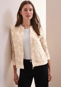 Chaqueta bomber beige con un patrón texturizado de hojas, puños acanalados y cremallera. Llevada sobre una camiseta blanca, combinada con pantalones negros.