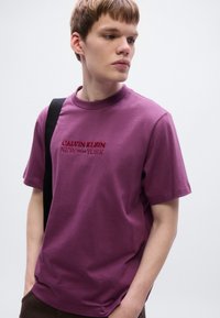 Κοντομάνικο μπλουζάκι με στρογγυλή λαιμόκοψη από βαμβάκι σε μωβ χρώμα μούρων, με κόκκινα κεντητά γράμματα "CALVIN KLEIN NEW YORK" στο μπροστινό μέρος. Μινιμαλιστικός σχεδιασμός.