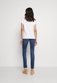 Vit kortärmad T-shirt i kombination med blå slim-fit jeans och beige plattformssandaler, sedd bakifrån mot en enkel bakgrund.