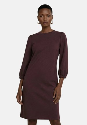 Femme portant une robe prune foncé jusqu'aux genoux, à manches longues avec des poignets élastiques et des boucles d'oreilles créoles, debout devant un fond clair uni.