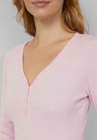 Jasnoróżowy top henley z długim rękawem, o prążkowanej fakturze, dekoltem w kształcie litery V i trzema guzikami na piersi. Prosty i dopasowany fason.