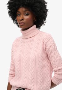 Pull à col roulé en maille torsadée rose clair avec col et poignets côtelés. Présente des motifs texturés et une coupe décontractée.