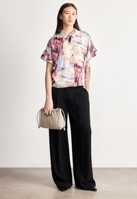 Žena v krátkém vícebarevném topu s krátkým rukávem, černých širokých kalhotách, držící béžovou plisovanou kabelku, stojící před jednoduchou zdí.