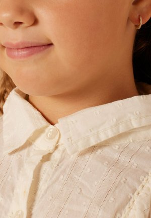 Witte blouse met kraag, gemaakt van een textuurstof met verhoogde stippen, verticale strepen en een knoopsluiting; kleine ringoorbel zichtbaar.
