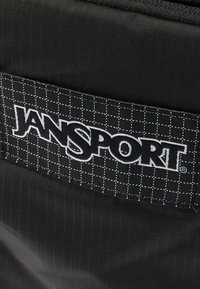 Μαύρο υφασμάτινο μπαλώμα με λευκές γραμμές πλέγματος και έντονο λευκό λογότυπο "JANSPORT" ραμμένο σε μαύρο υλικό με υφή.