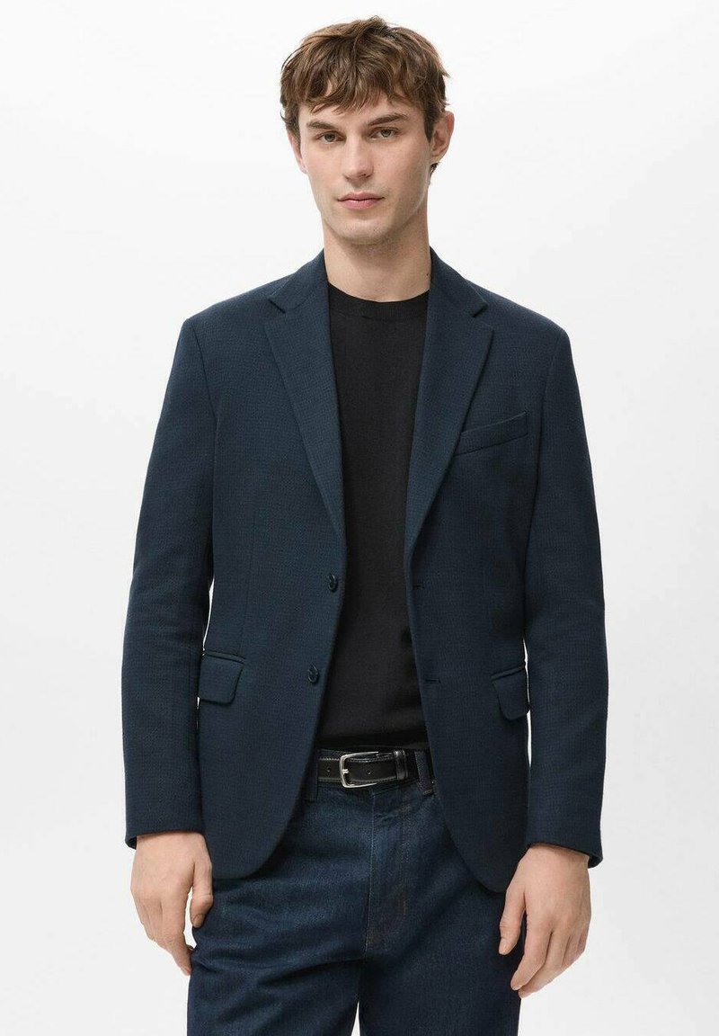 Donkerblauw colbert met een gestructureerde stof, enkelrijg ontwerp, revers met inkeping en een borstzak, gedragen over een zwart shirt en donkere spijkerbroek.