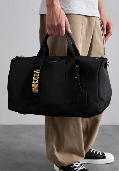 Black nylon duffle bag with gold hardware, featuring the brand name "Moschino" on the side, and a rectangular luggage tag attached.
