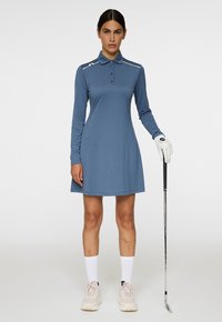 Nainen sinisessä pitkähihaisessa polo-mekossa ja golf-käsineessä pitelee golfmailaa, seisten yksinkertaista valkoista taustaa vasten.