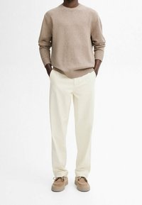 Homme portant un sweat-shirt beige à col rond, un pantalon droit blanc cassé et des chaussures décontractées beiges, debout les mains dans les poches sur un fond blanc.