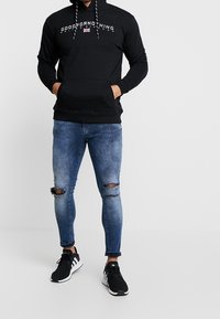 Schwarzer Hoodie mit "GOODFORNOTHING"-Aufschrift, Vordertasche und Kordelzügen. Kombiniert mit blauen, zerrissenen Slim-Fit-Jeans und schwarzen Sportschuhen.