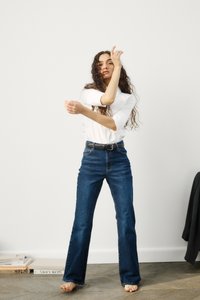 Bela srajca z zavihanimi rokavi, modri flared kavbojki z visokim pasom, črn pas, bosi, stoji ob običajnem ozadju z knjigami v bližini.