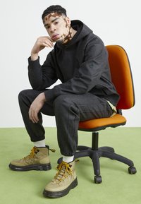 Sudadera y pantalones negros con textura, combinados con botas color beige que tienen cordones amarillos. Sentado en una silla naranja sobre un fondo de suelo verde.