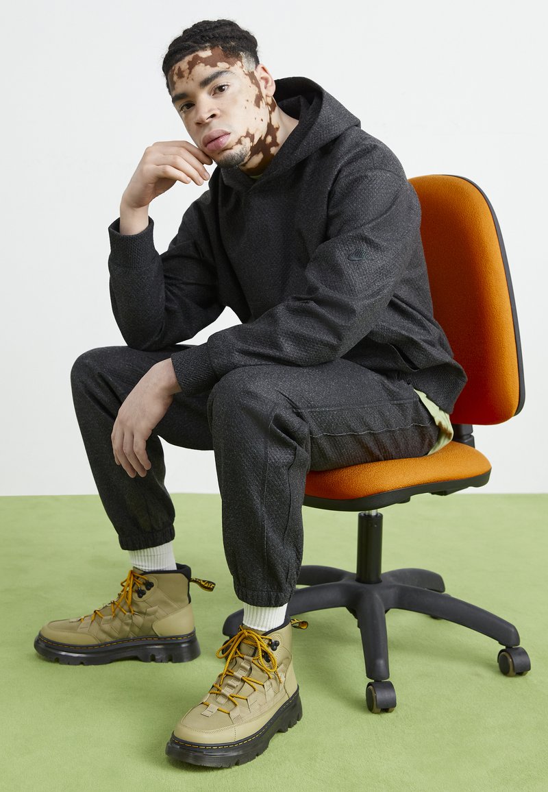 Sudadera y pantalones negros con textura, combinados con botas color beige que tienen cordones amarillos. Sentado en una silla naranja sobre un fondo de suelo verde.