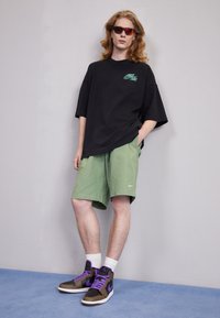Czarny oversize t-shirt, zielone szorty i brązowe trampki z purpurowymi akcentami. Model nosi czerwone okulary przeciwsłoneczne, stojąc na niebieskim dywaniku.
