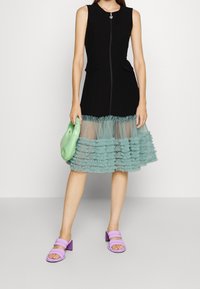 Robe noire sans manches avec une fermeture éclair à l'avant, ornée d'un ourlet en tulle vert menthe à volants. Tient une pochette verte, porte des sandales lilas à talons carrés.