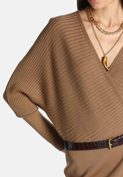 Femme portant un pull marron côtelé à col en V avec une ceinture foncée et des colliers en chaîne dorée superposés, dont un avec un pendentif en forme de corne.