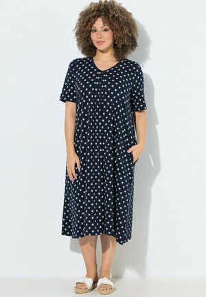 Frau mit lockigem Haar, die ein marineblaues Midi-Kleid mit weißen Sternmustern und weiße Schlupfsandalen trägt, posiert mit einer Hand in der Tasche.