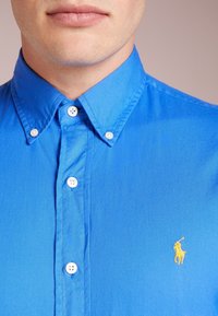 Blå skjorta i bomull med knapp, med krage, vita knappar och en gul broderad logotyp på bröstet. Slät textur.