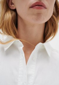 Balta apkakles krekls, kas izgatavots no viegla auduma, ar pogu aizdari priekšpusē un gludu tekstūru, ar redzamām šuvēm un apkakles detaļām.