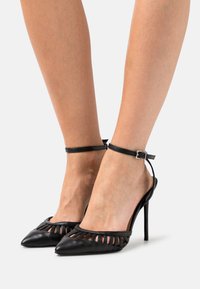 Sapatos de salto alto em couro preto com bico pontiagudo, laterais abertas e finas tiras no tornozelo. Apresentam detalhes recortados na parte superior.