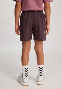 Barns mörklila träningsshorts med resårmidja, kombinerat med vita strumpor som har svarta chevronmönster och grå sneakers.