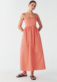 Abito maxi senza maniche di colore corallo con scollo a carré, vita arricciata e gonna fluida. Modello che indossa sandali neri piatti, su uno sfondo neutro.