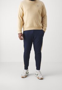 Sudadera beige con cuello redondo, combinada con pantalones deportivos azul marino con rayas laterales. Zapatillas blancas con detalles en verde y marrón.