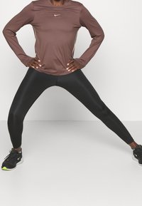 Brun långärmad träningsskjorta med rund halsringning och Nike-logotyp, tillsammans med svarta höga leggings och svarta sneakers med neonaccenter.