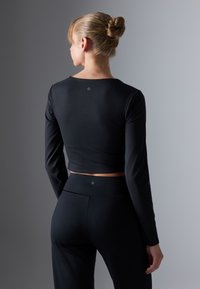 Žena s plavom kosom u punđi, obučena u crni dugih rukava crop top i crne hlače visokog struka, stoji naslonjena na pozadinu u dvije nijanse sive boje.