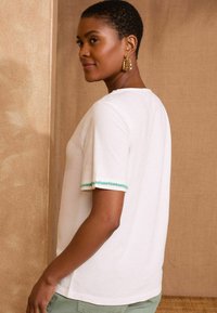 T-shirt blanche à manches courtes avec un col rond et des bordures en perles vertes sur les manches, fabriquée en tissu doux, présentée de côté sur un fond neutre.
