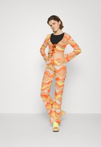 Conjunto de duas peças com padrão, apresentando uma parte de cima cropped de mangas longas com um decote profundo e cordão, combinado com calças flare em laranja e amarelo.